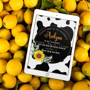 May include: A tablet displaying a birthday invitation with a black and white cow print background, a sunflower, and the text "Avelynn". The tablet rests on a bed of fresh yellow lemons.