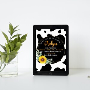 May include: A digital tablet displays a birthday invitation with a cow print background. The invitation features the name "Avelynn" in gold script, along with event details, a sunflower, and greenery. A glass vase with green leaves sits to the left.