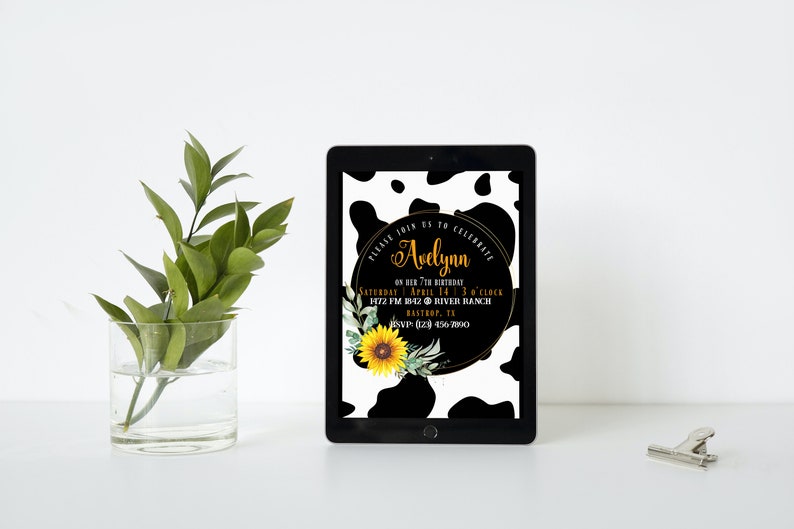May include: A digital tablet displays a birthday invitation with a cow print background. The invitation features the name "Avelynn" in gold script, along with event details, a sunflower, and greenery. A glass vase with green leaves sits to the left.