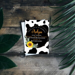 May include: A birthday invitation with a cow print design. The invitation features the name "Avelynn" in gold script, along with event details. A sunflower and greenery accent the design. The invitation is on a wooden background.