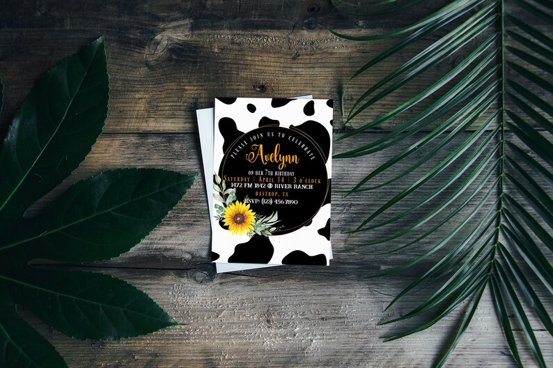 May include: A birthday invitation with a cow print design. The invitation features the name "Avelynn" in gold script, along with event details. A sunflower and greenery accent the design. The invitation is on a wooden background.
