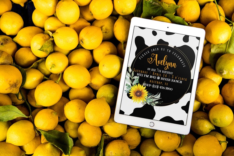 May include: A tablet displaying a birthday invitation with a black and white cow print background, a sunflower, and the text "Avelynn". The tablet rests on a bed of fresh yellow lemons.