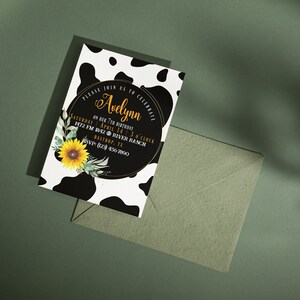 May include: A birthday invitation with a cow print background and a black circle with gold text that reads "Avelynn". The invitation includes a sunflower and greenery. An envelope is next to the invitation.