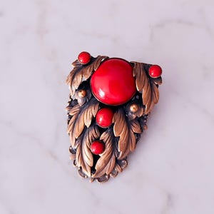 Peut inclure: Une broche ornée d'un grand cabochon rouge central entouré de cabochons rouges plus petits et d'un motif de feuilles en métal aux tons bronze. Le travail du métal présente des nervures complexes sur les feuilles, créant un aspect texturé autour des pierres rouges lisses et arrondies serties sur l'ensemble de la pièce.