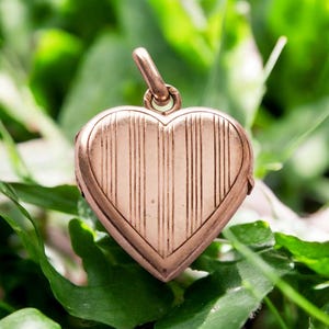 Op de afbeelding: Een hartvormige medaillonhanger van metaal in rozetinten, voorzien van een reeks verticale gegraveerde lijnen over het oppervlak en een kleine ronde ring aan de bovenkant voor bevestiging aan een ketting of koord in een eenvoudig ontwerp.