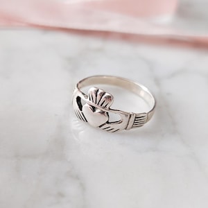 May include: A silver Claddagh ring, a traditional Irish design, featuring a heart, crown, and hands. The ring sits on a white marble surface, with a pink ribbon in the background. The ring is a symbol of love, loyalty, and friendship.