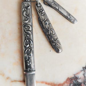 May include: A set of three antique silver-coloured items, including a pencil holder and two small knives, linked by a ring. Each piece features an elaborate floral and leaf design. The items are presented on a light-coloured, marble-effect surface.
