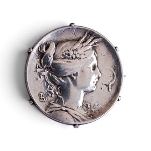 Peut inclure: Une broche circulaire en argent présentant le profil en relief d'une personne portant une couronne de blé dans les cheveux. La surface métallique présente une finition ternie avec des détails gravés, dont un monogramme en bas à gauche.