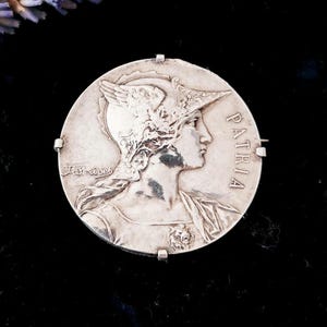May include: Silver toned brooch featuring a profile of a woman wearing a helmet with wings. The brooch is round and has the word "PATRIA" inscribed on the right side.