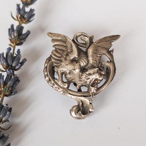 May include: A silver-toned brooch featuring a mythical griffin with outstretched wings and a dragon-like head. The brooch has intricate detailing and is set against a white background with a sprig of lavender.