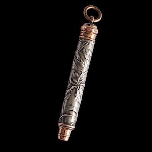 May include: A silver-colored, cylindrical object with intricate floral engravings. The top and bottom are accented with copper-colored metal. A small copper-colored ring is attached to the top. The object is set against a black background.