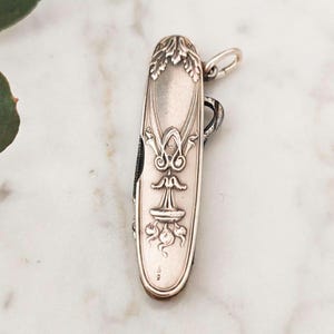 May include: A silver pocket knife with an ornate floral design and a loop for attaching a chain. The knife is closed and has a maker's mark on the back.