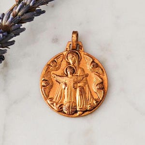 May include: A gold-toned metal pendant featuring a detailed relief of the Virgin Mary holding a baby Jesus. The pendant is surrounded by angels with wings and is a religious symbol.