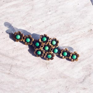 Peut inclure: Une barrette à cheveux composée d'un groupe de fleurs en métal bronze, chacune ornée en son centre d'une petite pierre cabochon ronde vert vif. L'arrangement floral est allongé, les fleurs se chevauchant pour former une pièce unique.