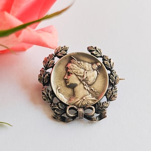 Peut inclure: Broche argentée représentant un profil classique de femme dans un cadre circulaire, entouré d'une couronne de laurier et d'un nœud. La broche est probablement vintage ou antique.