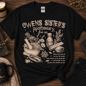 May include: A black t-shirt featuring a beige graphic print of a mortar and pestle, herbs, a bottle, and cinnamon sticks. The text reads: Owens Sister's Apothecary est. 1995, along with a quote about salt, rosemary, lavender, and falling in love.