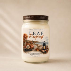 May include: A 12oz soy blend candle in a clear glass jar with a brown lid. The label features an illustration of an orange truck, a mug, and a lantern by a lake, with the text THE CABIN BOUTIQUE CO, LEAF PEEPING, 12oz 340g Soy Blend Candle, Made in Connecticut.
