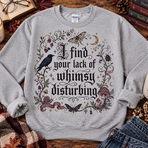 以下が含まれることがあります： I find your lack of whimsy disturbingという黒いグラフィックプリントが施されたライトグレーのクルーネックトレーナー。テキストの周囲には、カラス、蛾、蝶、カタツムリ、甲虫、キノコが描かれた詳細なイラストが配置されています。