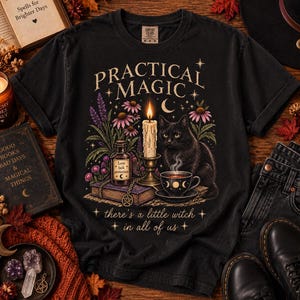 May include: A black t-shirt featuring a graphic print with the text "PRACTICAL MAGIC" and "there's a little witch in all of us". The design includes a black cat, a lit candle, a teacup, a potion bottle, a book, and purple and pink flowers.