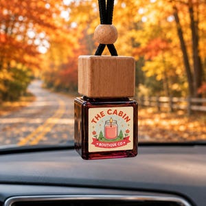 Pode incluir: Um frasco quadrado de vidro para ambientador de carro com tampa de madeira e cordão preto. O rótulo apresenta um gráfico de uma vela acesa num frasco, dois pinheiros e o texto THE CABIN BOUTIQUE CO em letras vermelhas e cor de laranja.