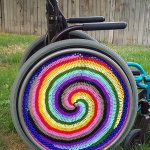 Könnte beinhalten: Ein Rollstuhl mit einer bunten gehäkelten Radabdeckung. Die Abdeckung ist ein Regenbogen-Spiralmuster mit einer schwarzen Mitte.