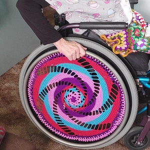 Op de afbeelding: Een rolstoel met een kleurrijke gehaakte wielbeschermer. De beschermer is een spiraalvormig patroon in roze, paars, blauw en rood.