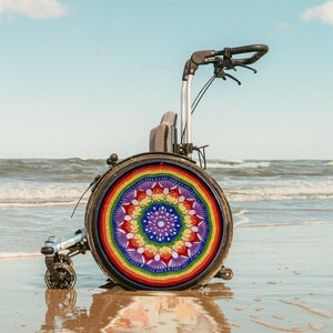 Op de afbeelding: Een rolstoel met een kleurrijk gehaakt mandala-ontwerp op het achterwiel. De rolstoel staat op een zandstrand met blauw water op de achtergrond.