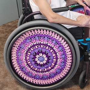 Pode incluir: Uma cadeira de rodas com uma capa de crochê em tons de rosa e roxo. A capa é um design de mandala com um círculo central e várias camadas de detalhes de crochê.