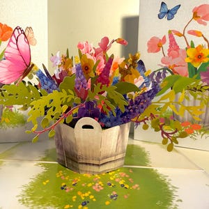May include: A 3D pop-up card with a wooden bucket filled with colourful paper flowers. Pink butterflies and other flowers are also present. The card is open, revealing a grassy scene with more flowers.