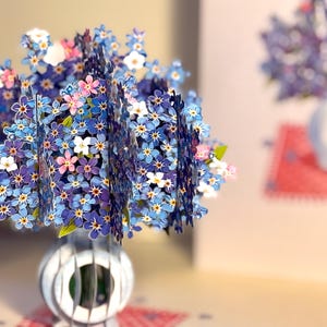 May include: A bouquet of blue forget-me-not flowers with a few pink flowers in a white vase. The flowers are arranged in a circular shape and the vase is made of a white material.