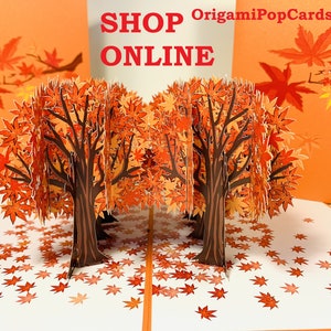 May include: A 3D pop-up card featuring two trees with orange and red leaves. The trees are surrounded by scattered leaves on a white background. The card is open and the trees are standing upright. The text "SHOP ONLINE" and "OrigamiPopCards.com" is printed on the card.