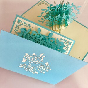 Puede incluir: Tarjeta de felicitación desplegable azul claro con diseño floral. La tarjeta presenta flores y hojas turquesas cortadas con láser, con un diseño floral a juego en la parte delantera. El interior revela un arreglo floral tridimensional.