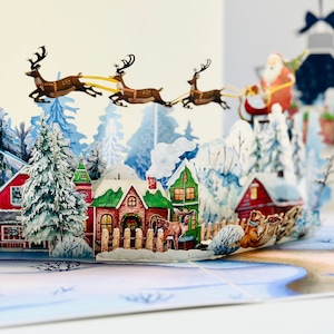May include: A pop-up Christmas card featuring a winter village scene with snow-covered houses, evergreen trees, and Santa Claus in a sleigh pulled by reindeer. The card has a three-dimensional design with intricate details and a festive holiday theme.