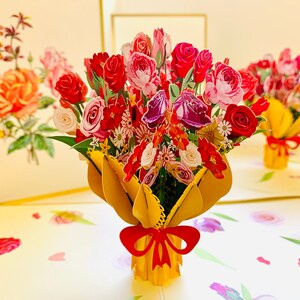 May include: A 3D pop-up card featuring a bouquet of red, pink, and purple flowers in a yellow paper wrapper with a red bow. The card is open and the flowers are standing up.