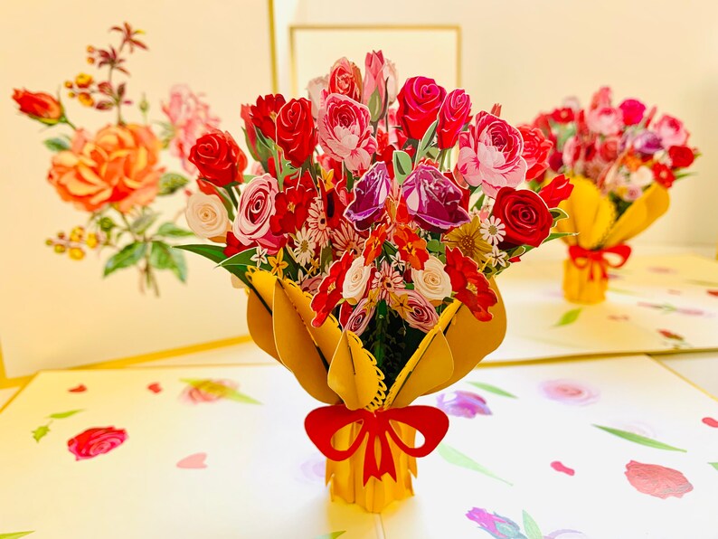 May include: A 3D pop-up card featuring a bouquet of red, pink, and purple flowers in a yellow paper wrapper with a red bow. The card is open and the flowers are standing up.