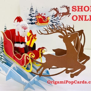May include: A 3D pop-up Christmas card featuring Santa Claus in a red sleigh pulled by reindeer. The card is decorated with snow-covered trees and a snowy landscape. The text "SHOP ONLINE" is printed on the card. The website address "OrigamiPopCards.com" is printed at the bottom of the card.