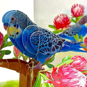 May include: A detailed 3D paper art piece depicting two blue parakeets on a brown branch with pink and red flowers. The parakeets have intricate feather patterns and yellow markings. The background is white, creating a striking contrast.