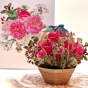 May include: A 3D pop-up greeting card featuring a basket of pink flowers with a blue ribbon. The card is set against a backdrop of a floral illustration with pink roses and white flowers. The basket is made of woven material.