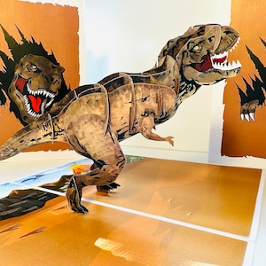 Puede incluir: Una tarjeta emergente 3D con un dinosaurio Tyrannosaurus Rex. El dinosaurio marrón y beige rompe un fondo marrón, con una escena de paisaje detallada en la base. El T-Rex tiene boca roja y dientes blancos.