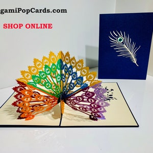 May include: A rainbow peacock pop-up card with a blue envelope. The card is open and the peacock's tail feathers are spread out. The feathers are made of paper and are cut out in the shape of peacock eyes. The envelope has a single white peacock feather with a blue eye on it.