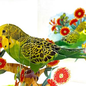 May include: A 3D paper art piece featuring two budgerigars perched on a branch with red and yellow flowers. The birds have yellow heads, green bodies, and black markings. The artwork is set against a white background.