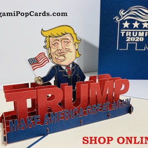 May include: A red, white, and blue pop-up card featuring a cartoon depiction of Donald Trump holding an American flag. The card reads "TRUMP MAKE AMERICA GREAT AGAIN".