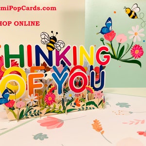 May include: A colorful 3D pop-up card with the words "Thinking of You" in a rainbow of colors. The card features a bee, butterflies, and flowers.