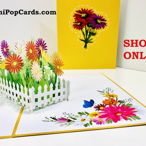May include: A 3D pop-up card featuring a white picket fence surrounding a garden with colourful flowers. The card is open and the flowers are popping up. The card is on a white surface with a yellow background. The text "OrigamiPopCards.com" is in the top left corner of the image. The text "SHOP ONLINE" is in the top right corner of the image.