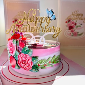May include: A pop-up anniversary card featuring a pink cake decorated with red roses and green leaves. The cake has a gold glitter "Happy Anniversary" topper and a blue butterfly. A matching card is in the background.