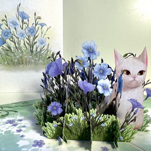 May include: A 3D pop-up card with a white cat surrounded by blue flowers and green leaves. The card's design includes detailed paper cutouts of the cat and floral elements. A watercolor painting of blue flowers is on the left.