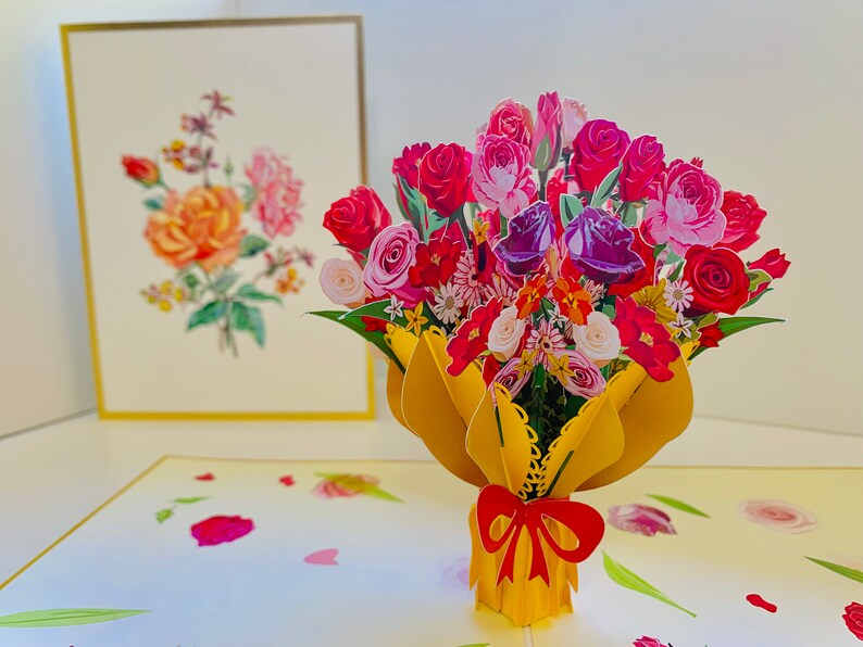 May include: A 3D pop-up card featuring a bouquet of colorful roses, lilies, and other flowers in a yellow vase with a red bow. The card is open and the flowers are standing up.