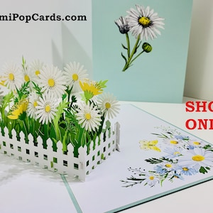 May include: A 3D pop-up card featuring a white picket fence surrounding a garden of white daisies and green grass. The card is open and the daisies are popping up. The card is on a white background with a blue background behind it. The text "SHOP ONLINE" is on the blue background.