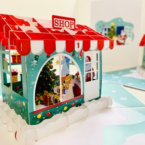 Puede incluir: Una tarjeta de Navidad en 3D con un pop-up que representa una tienda con rayas rojas y blancas y un toldo verde. La tienda tiene una ventana con un árbol de Navidad, regalos y una figura de Papá Noel en su interior. La tarjeta está decorada con copos de nieve y luces.