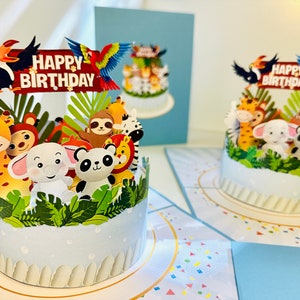 Puede incluir: Tarjetas de cumpleaños emergentes con un diseño de pastel blanco, animales de la jungla y las palabras "Happy Birthday". Las tarjetas tienen un tucán y un loro en la parte superior. Los animales incluyen una jirafa, un mono, un perezoso, una cebra, un león, un elefante y un panda.